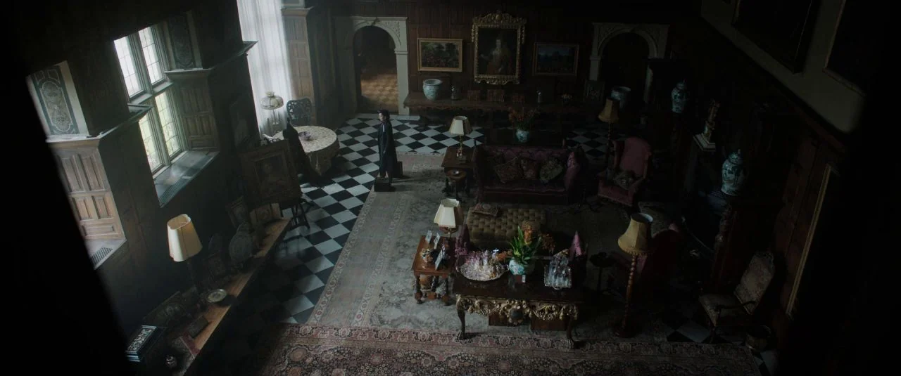 Rebecca movie still: wide — A dark, wood-paneled room features a black-and-white checkered floor and ornate period furniture. A lone person in a…