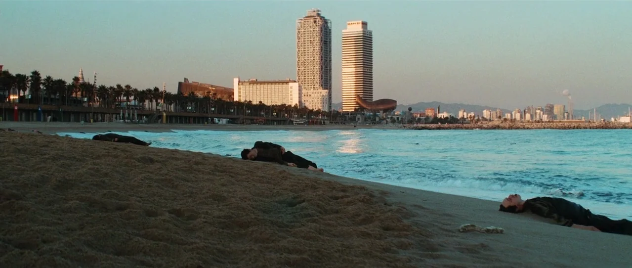 Biutiful (2010) movie still: wide — A sandy beach stretches toward a city skyline featuring prominent twin skyscrapers. Three men lie facedown or on…
