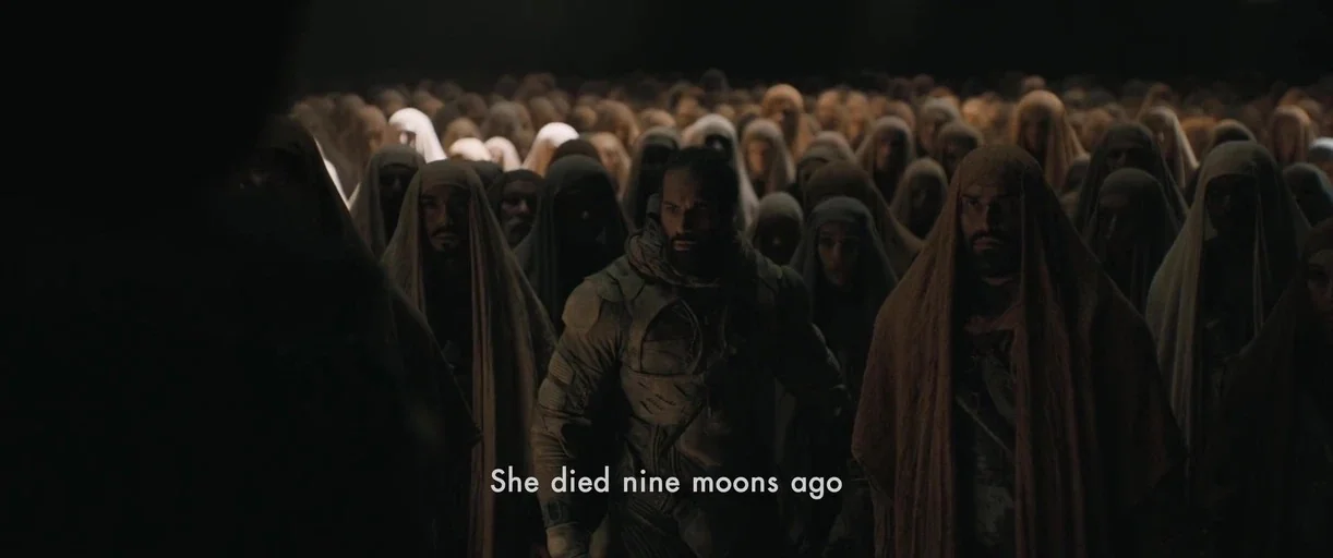 Dune: Part 2 (2024) movie still: wide — A dense gathering of people wearing robes stands in a dark desert environment. The figures look toward the…