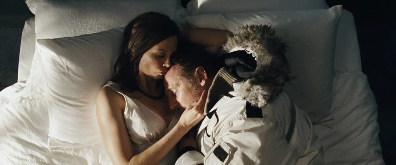 The Grey (2012) movie still: medium — A man in a winter parka and a woman in a white top lie in bed. The woman kisses the forehead of the man while…
