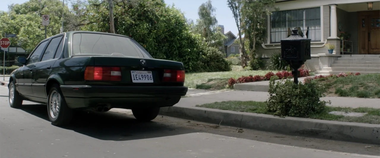 Truth or Dare (2018) movie still: medium — A suburban street features a dark green sedan parked near a roadside mailbox. The background shows a…