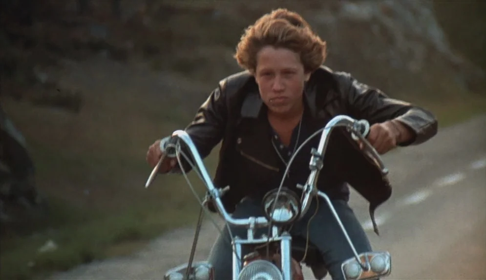A Swedish Love Story (1970) movie still: medium — A paved country road stretches out behind a lone motorcyclist in motion. The rider balances on the…