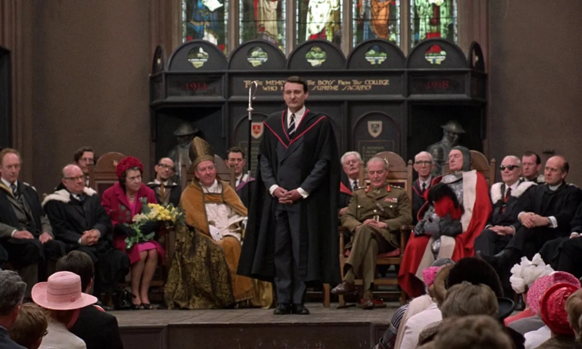 If…. (1968) movie still: medium — A formal hall features a dark wooden memorial screen behind a raised wooden stage. A man stands at the front,…