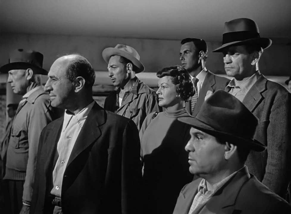 M (1951) (1951) movie still: medium — A group of six people stand close together in a dimly lit interior space. They appear to be waiting or…
