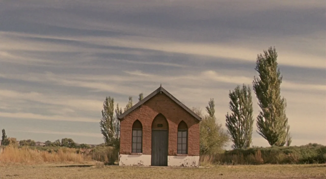 Patagonia (2010) movie still: wide — A small brick building with pointed arched windows sits alone in a flat landscape. Tall, thin poplar trees…