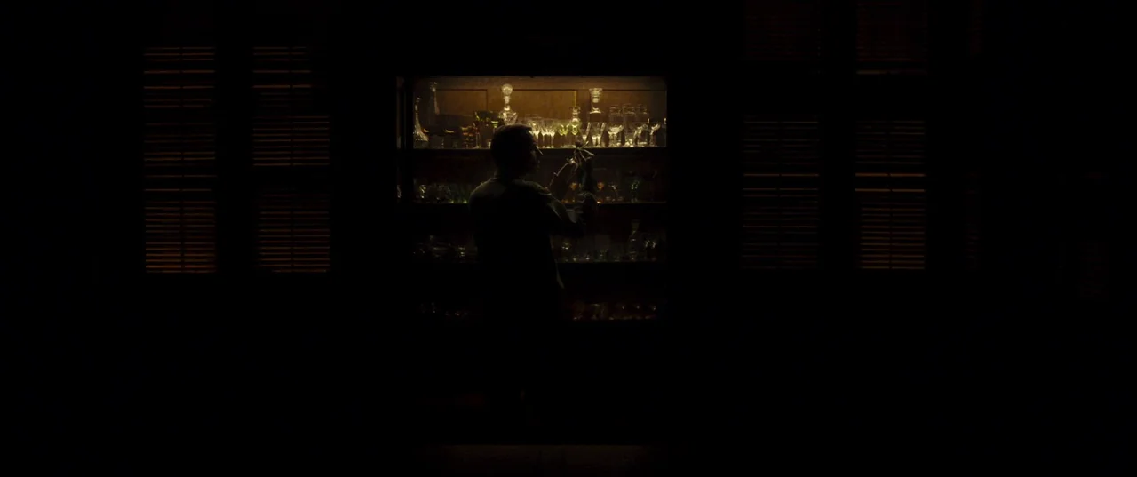 Earwig (2022) movie still: medium — A dimly lit room features a large glass cabinet filled with crystal glassware. A man stands in front of the…