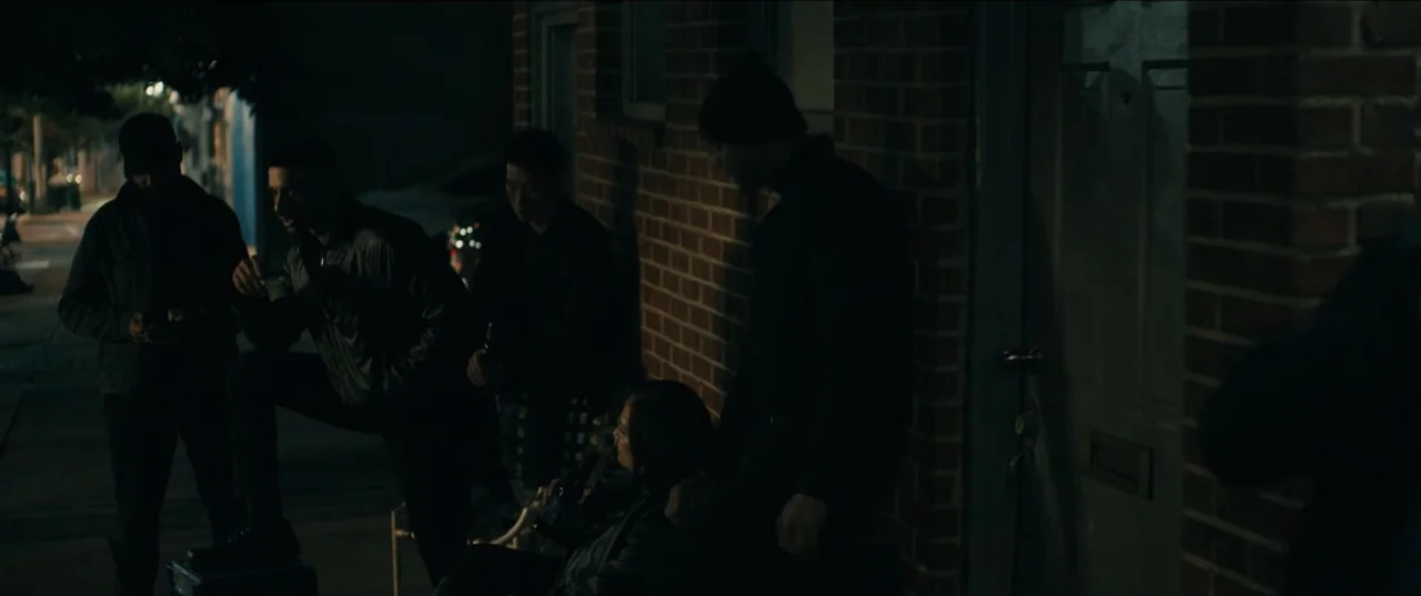 Guns Up (2025) movie still: medium — A group stands against a dark red brick wall by a closed door. They are gathered outside at night under dim…