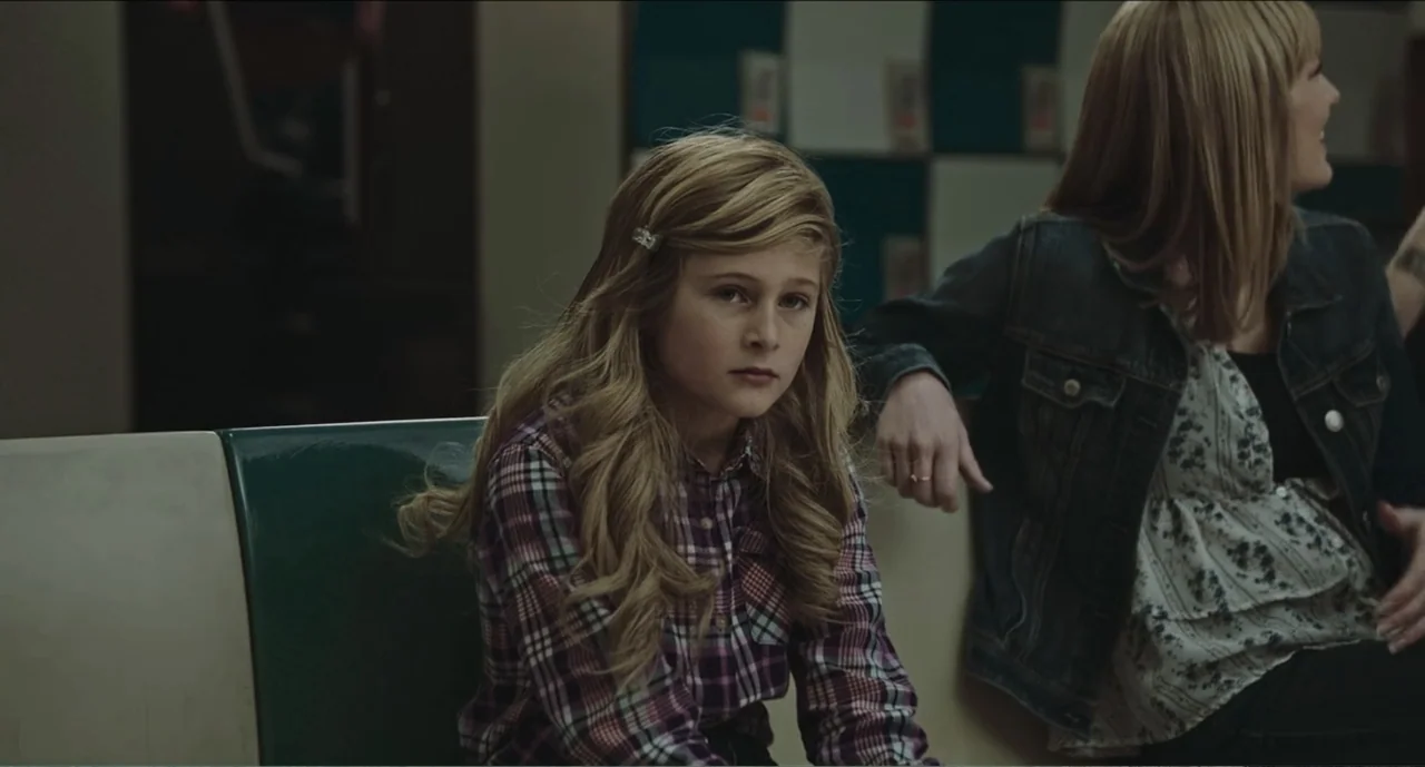 Cowboys (2014) movie still: medium — A interior view of a retro diner booth with padded green vinyl seating. A young girl in a plaid shirt sits…