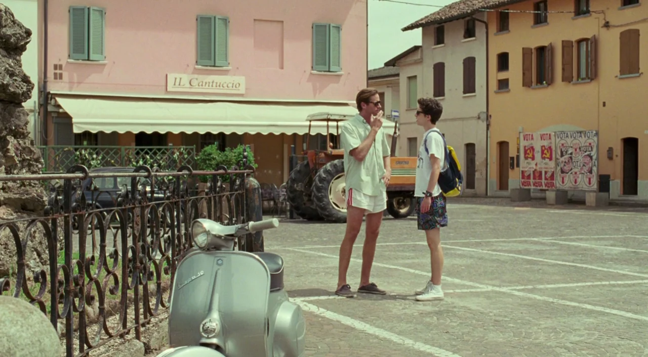 Call Me By Your Name (2017) movie still: wide — A sunny Italian town square features two men conversing near a parked vintage scooter. Stucco…