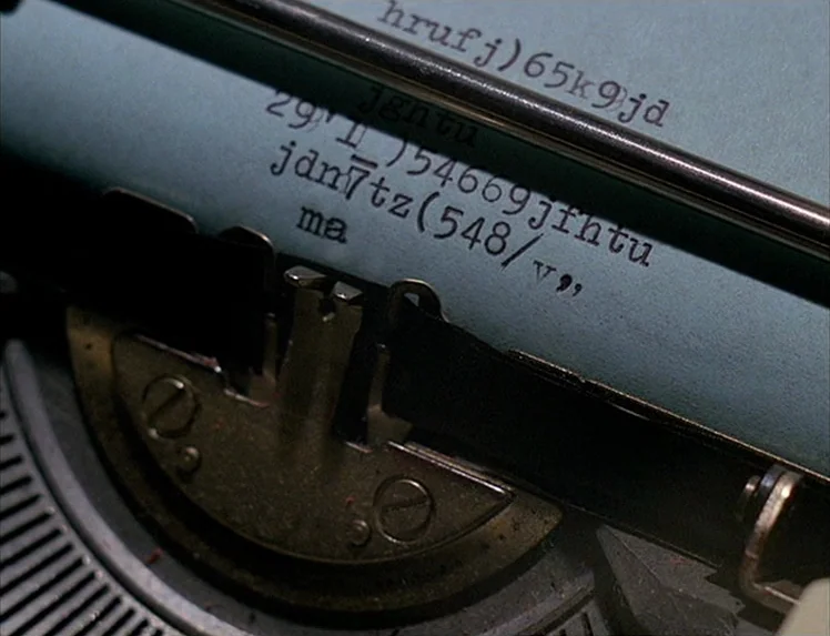 First Name: Carmen (1983) movie still: closeup — A close-up of a vintage typewriter platen holding a blue sheet of paper. The paper contains…