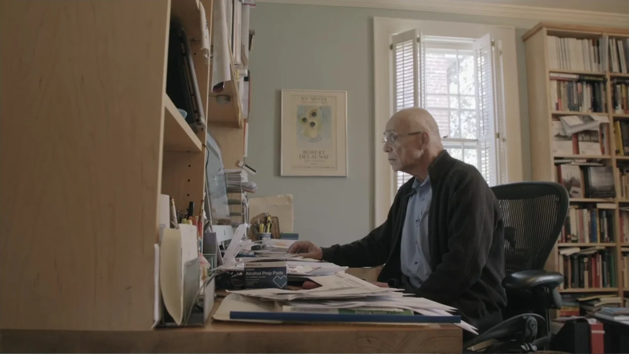 A Gift from God (2019) movie still: medium — An older man sits at a desk cluttered with papers in a home office. He looks at a desktop computer…