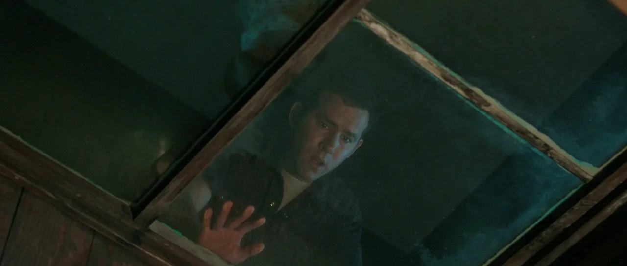 The Voices (2014) movie still: medium — A dark, cramped space with wooden paneling and a thick glass overhead barrier. A man looks up through the…