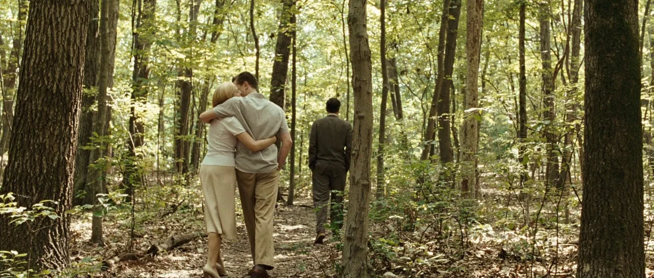 Revolutionary Road (2008) movie still: fullbody — A dense forest path with tall deciduous trees and scattered dry leaves on the ground. A couple…