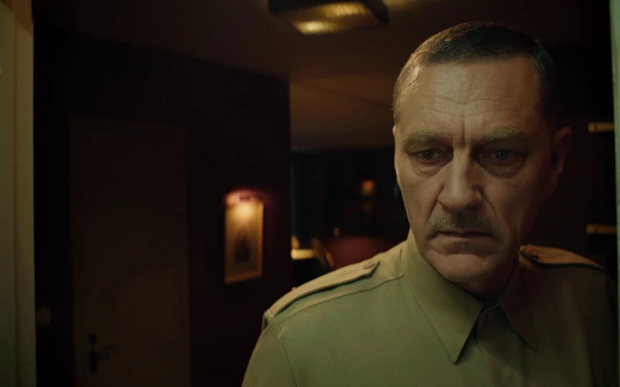 Knocking (2021) movie still: closeup — A dimly lit indoor corridor with dark walls and a soft light source. An older man in military-style clothing…