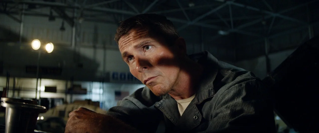 Ford V Ferrari (2019) movie still: medium — A dark automotive garage with exposed rafters and distant indistinct industrial equipment. A man in a…