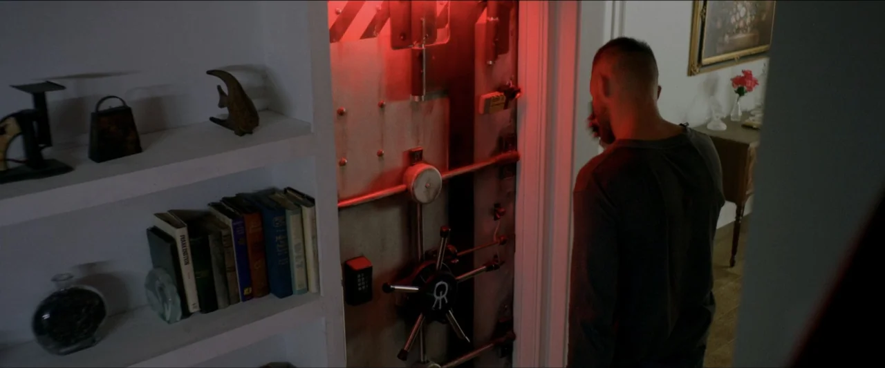 Ambush (2020) movie still: medium — A dimly lit residential study features built-in white shelves filled with books and decorations. A man in a dark…