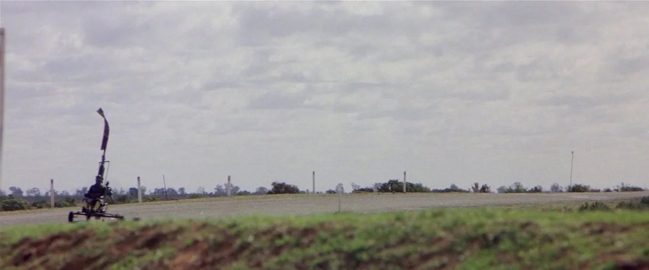 Mad Max 2 (1981) movie still: wide — A flat asphalt road stretches across an open, overcast rural landscape. A small three-wheeled vehicle with a…