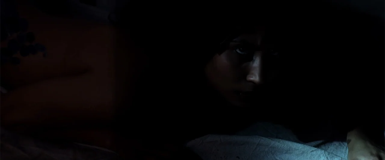 Mermaid Down (2019) movie still: closeup — A dimly lit room features a person reclining on white sheets in deep shadow. A young woman with dark hair…