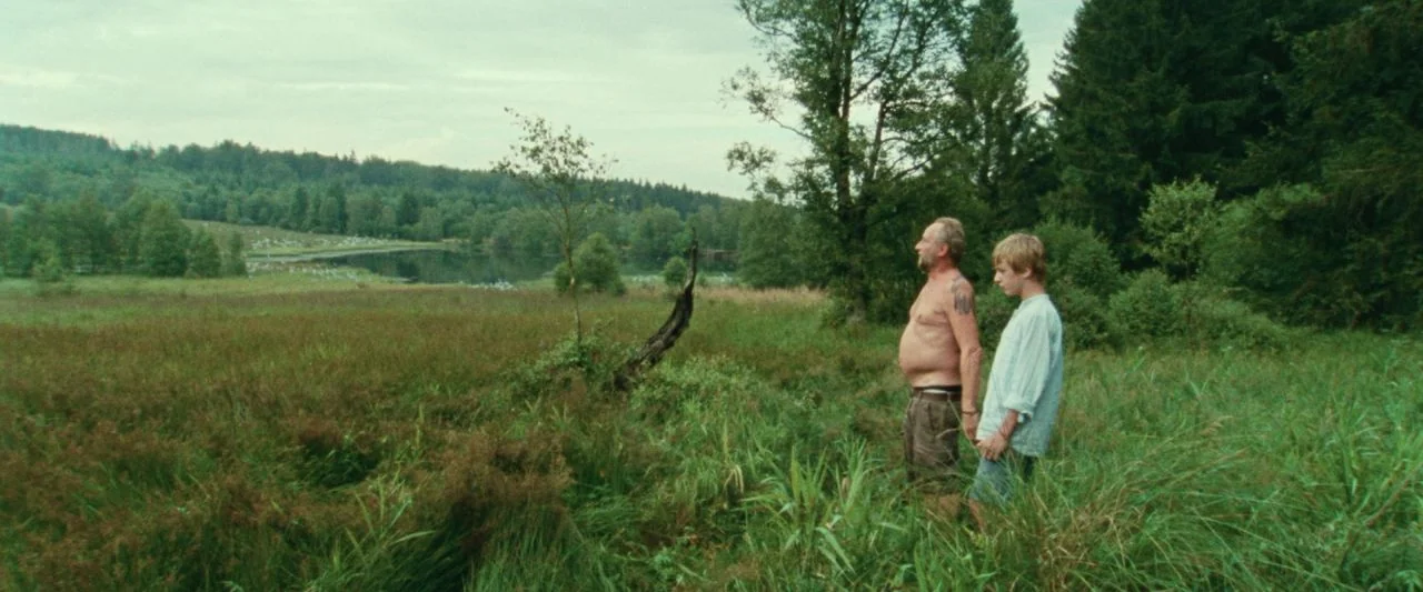Adoration (2019) movie still: wide — An open grassy landscape features a small lake and dense surrounding forest background. An older man and a…