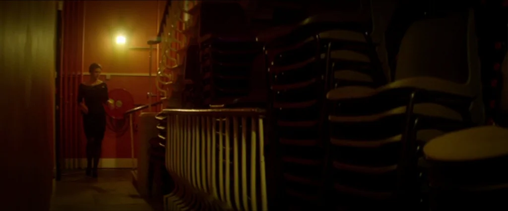 Haywire (2011) movie still: wide — A narrow corridor leads past stacks of chairs in a dimly lit theater space. A woman in a black dress walks slowly…