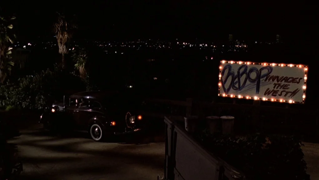 Bird (1988) movie still: wide — A dark hillside area at night features a car parked next to a billboard. The billboard features illuminated bulbs…