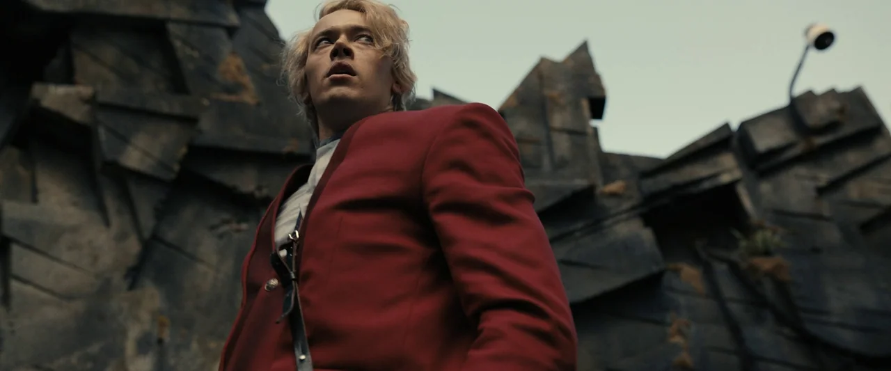 The Hunger Games: The Ballad of Songbirds & Snakes (2023) movie still: medium — A man in a red blazer stands before a backdrop of jagged concrete…
