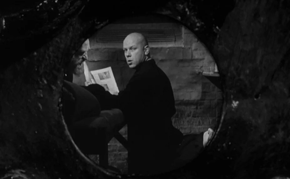The Criminal (1960) movie still: medium — Two men sit inside a stone cell viewed through a small, jagged circular aperture. The bald man looks…