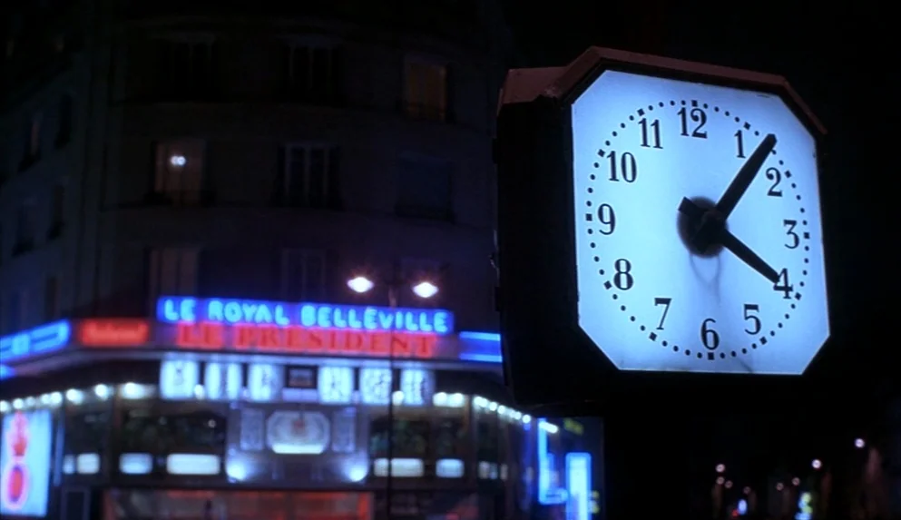Night on Earth (1991) movie still: closeup — A glowing outdoor clock stands in the foreground of a dark city street intersection. The background…