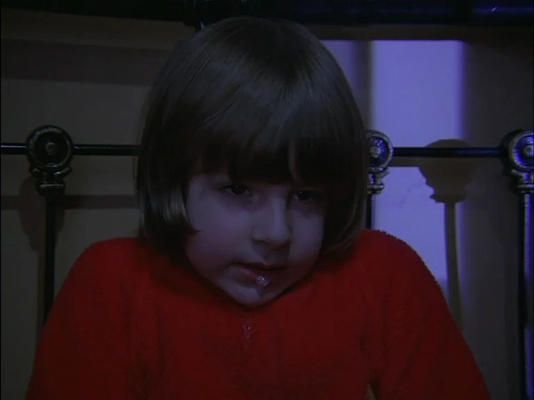 The Shining (1980) movie still: closeup — A dimly lit indoor space with a dark metal bed frame structure visible behind. A young child with short…