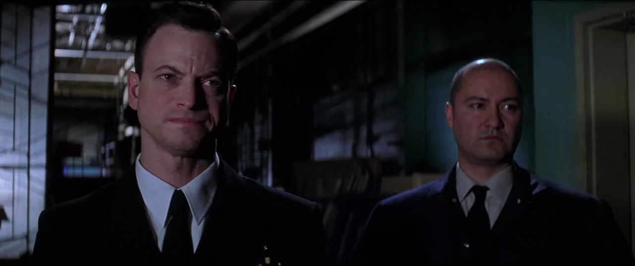 Snake Eyes (1998) movie still: medium — A dimly lit narrow hallway with indistinct industrial architectural details in the background. Two men in…