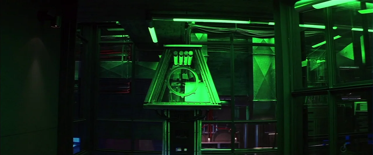 Mission: Impossible II (2000) movie still: establishing — A dark, industrial laboratory features exposed wiring, ventilation ducts, and a central…