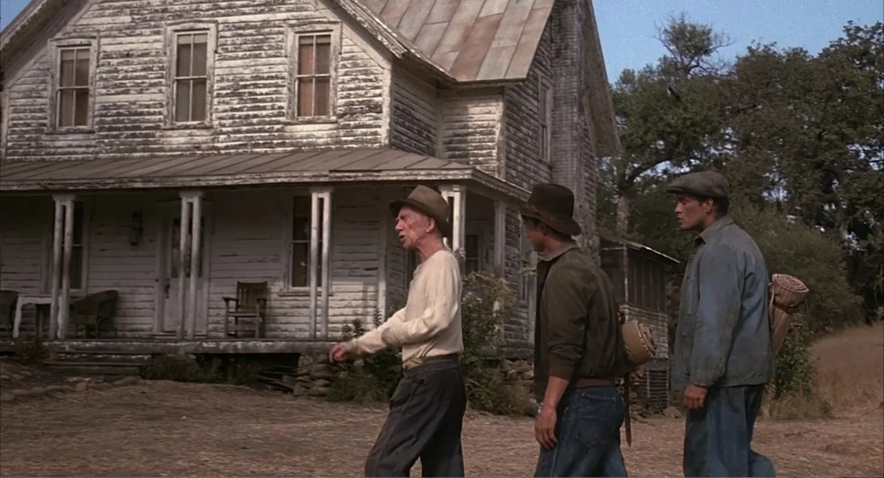 Of Mice and Men (1992) movie still: fullbody — A weathered two-story white farmhouse stands behind a group of three walking men. The three men walk…