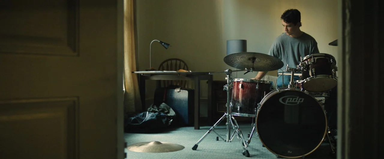 Whiplash (2014) movie still: medium — A sparsely furnished room features a drum kit placed prominently in the foreground. A young man sits behind…
