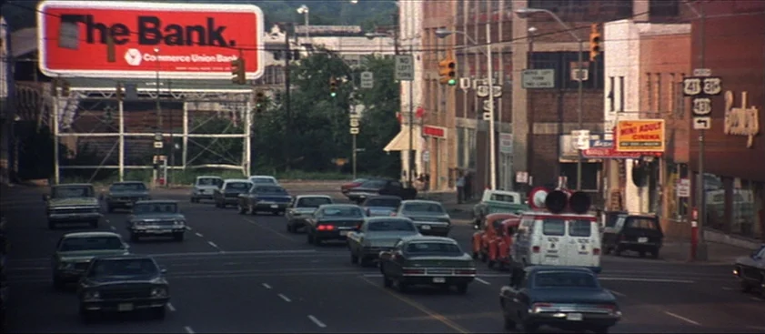 Nashville (1975) movie still: wide — A city intersection in the 1970s features a large red bank billboard overhead. Multiple vintage cars wait in…