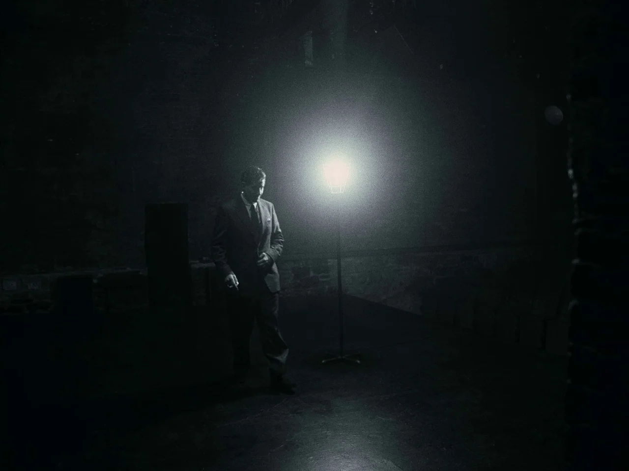 Maestro (2023) movie still: fullbody — A dark, sparse room with a stone wall and an exposed lightbulb stand. A man in a suit walks across the floor…