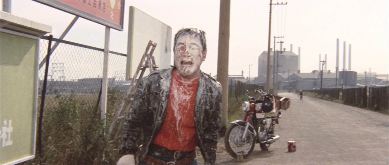All Monsters Attack (1969) movie still: medium — An industrial landscape features a dirt road lined with chain-link fences and utility poles. A man…