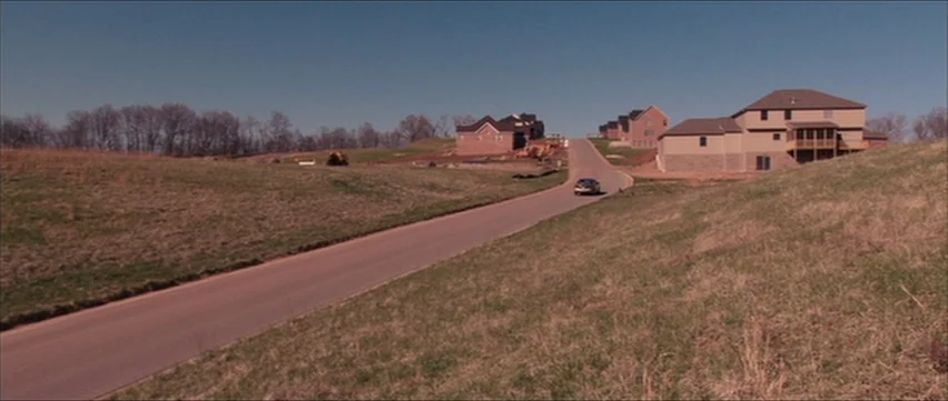 Bubble (2005) movie still: wide — A winding asphalt road leads through undeveloped rolling hills toward a small suburban development. A single small…