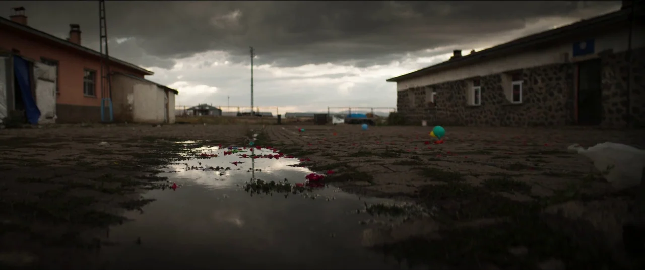 About Dry Grasses (2023) movie still: establishing — An overcast paved square sits between two low-slung, rustic buildings under dark storm clouds.…