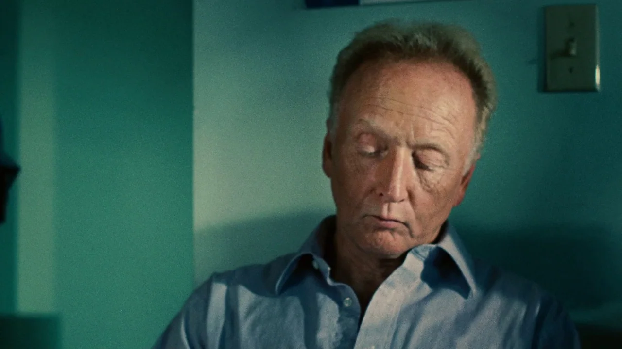Saw II movie still: closeup — A dimly lit room with teal-painted walls and a light switch on the wall. An elderly man with light hair sits with his…