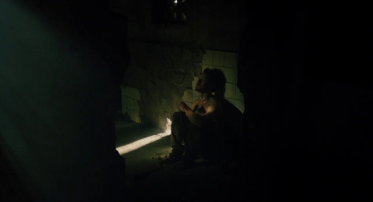 Submergence movie still: fullbody — A dark concrete enclosure with crumbling white tiles on the rear wall. A shirtless man sits on the floor inside…