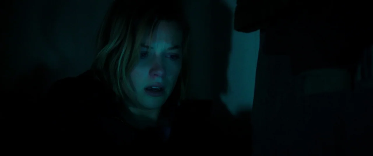 Don’t Breathe (2016) movie still: closeup — A dimly lit room with heavy shadows casting across the surrounding environment. A young woman stares at…
