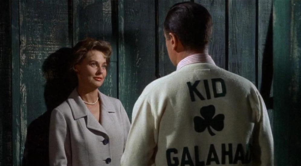 Kid Galahad (1962) movie still: medium — A man and woman talk in front of a dark green wooden wall. The man wears a cream cardigan displaying his…