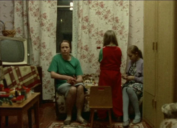 From The East (1993) movie still: medium — A domestic space with floral wallpaper features a television and wooden furniture. A woman sits on a…