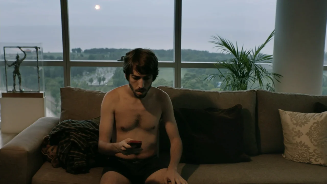 That's Not Cheating (2016) movie still: medium — A modern apartment with a large window overlooking a landscape at daytime. A shirtless man sits on…
