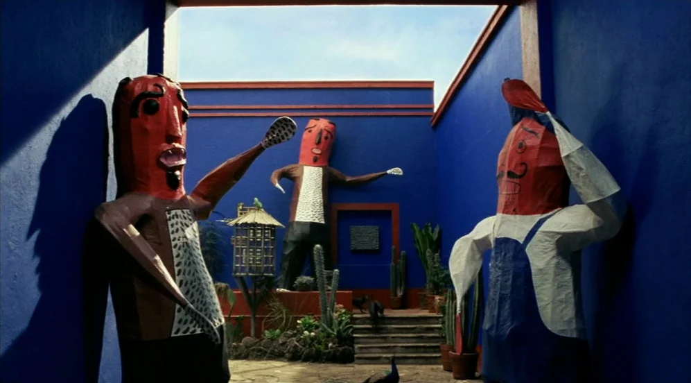 Frida (2002) movie still: wide — A bright blue courtyard contains three large, stylized papier-mache figures and several potted cacti. The figures…