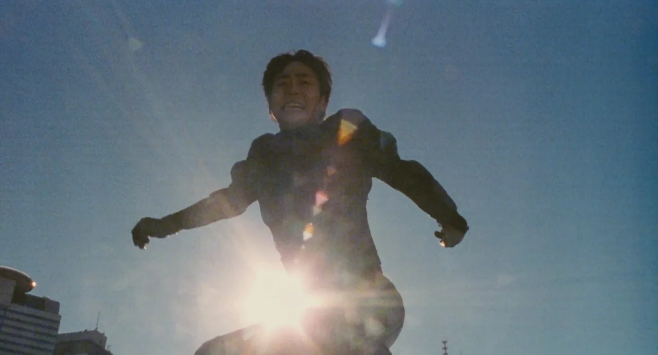 Ichi The Killer (2001) movie still: medium — An urban cityscape is visible at the very bottom edge of the bright sky. A man in dark clothing leaps…