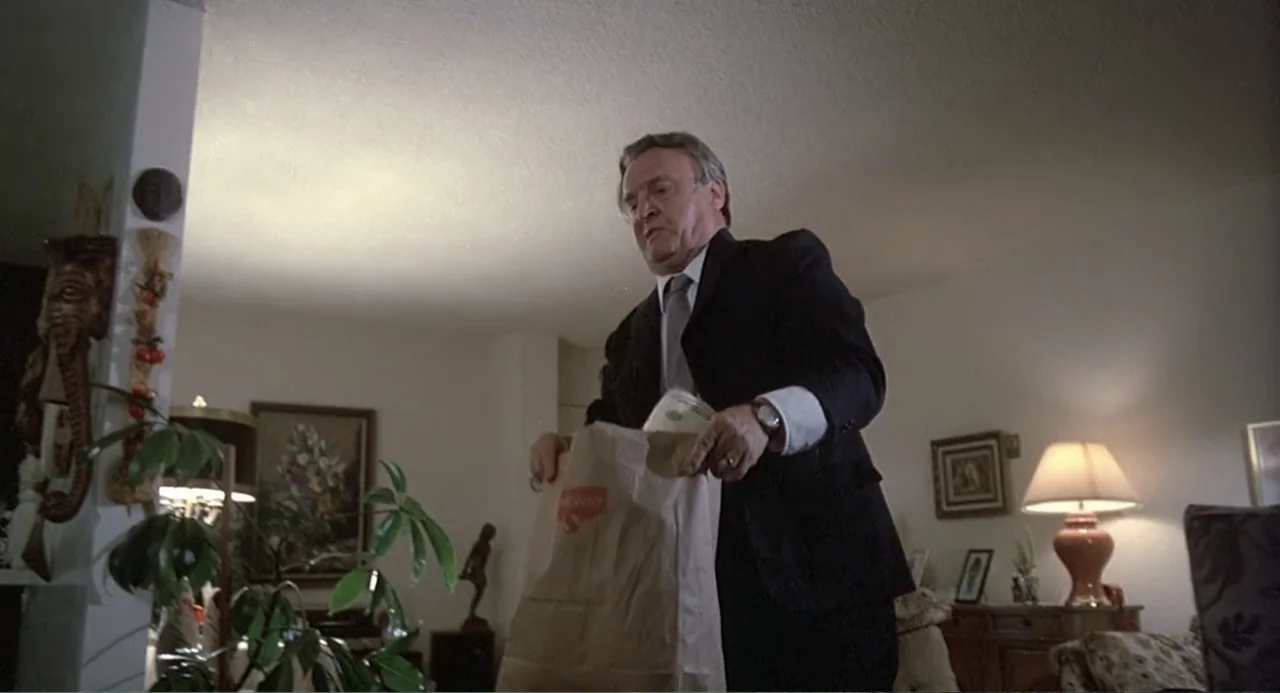 Death Wish II (1982) movie still: medium — A furnished living room features white walls, hanging decor, and a table lamp. An older man in a business…