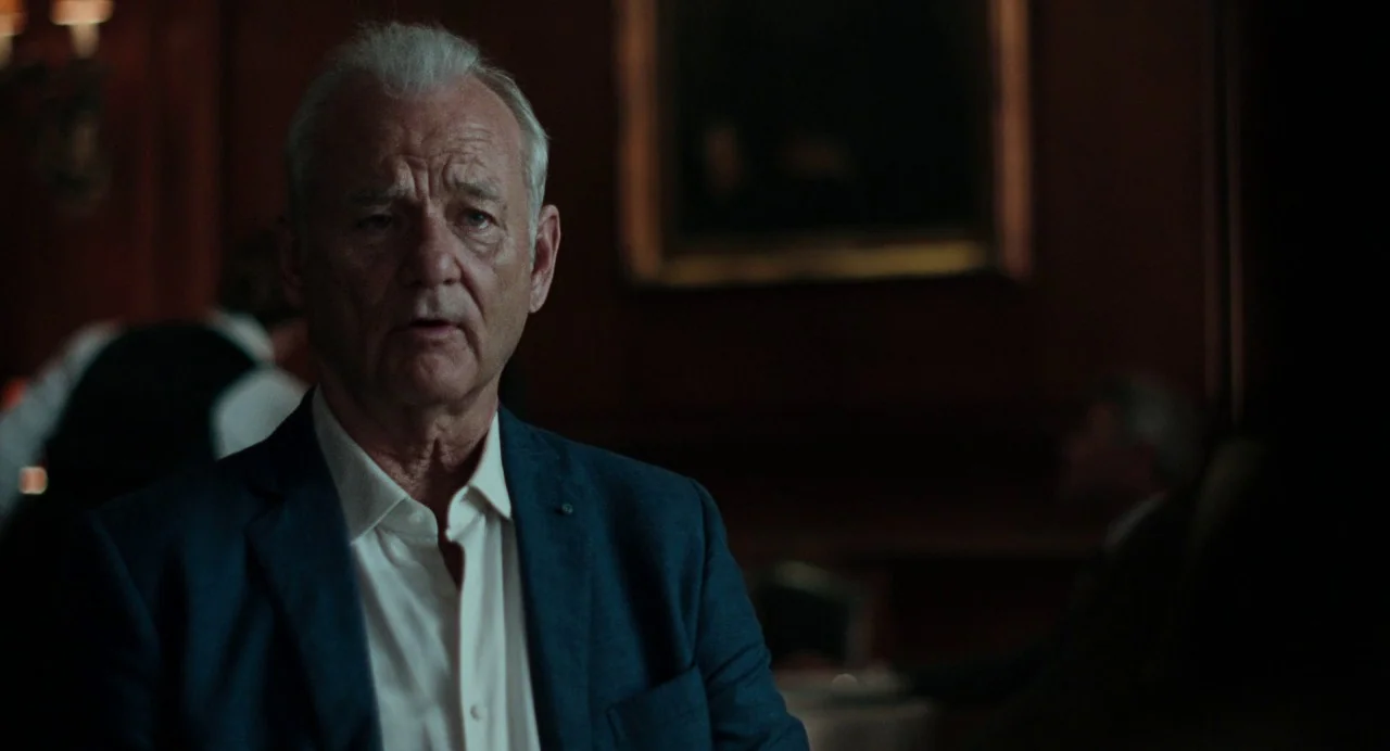 On The Rocks (2020) movie still: medium — A dimly lit room with dark wood paneled walls and a blurred framed picture. An older man in a blue blazer…