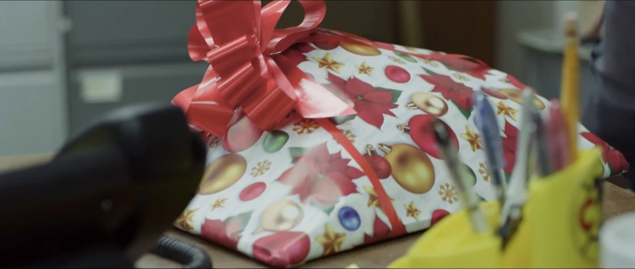 Mirreyes vs Godínez (2019) movie still: closeup — A desk surface holds a gift wrapped in red, white, and gold holiday paper. A yellow pen holder…