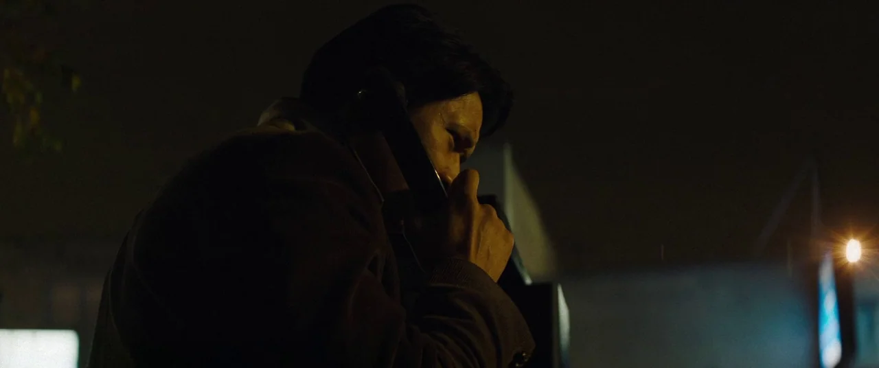 Broadcast Signal Intrusion (2021) movie still: closeup — An exterior nighttime street environment at a phone booth. A man stands in the dark while…